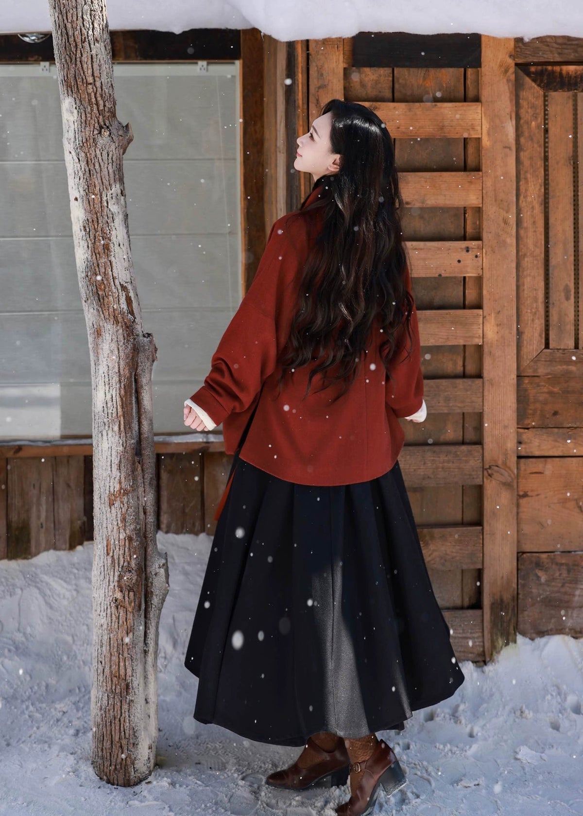 Woman in a red coat and black skirt standing in the snow next to a wooden structure.